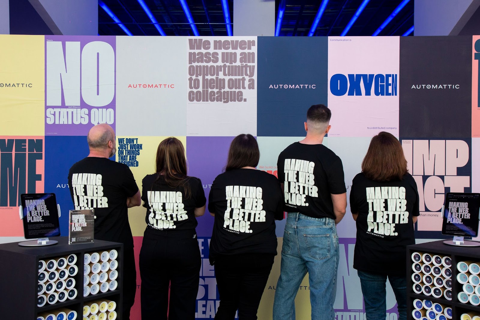 Group of Automattic employees wearing black shirts with the phrase 'Making the web a better place' on the back, standing in front of a colorful wall with various slogans related to Automattic.