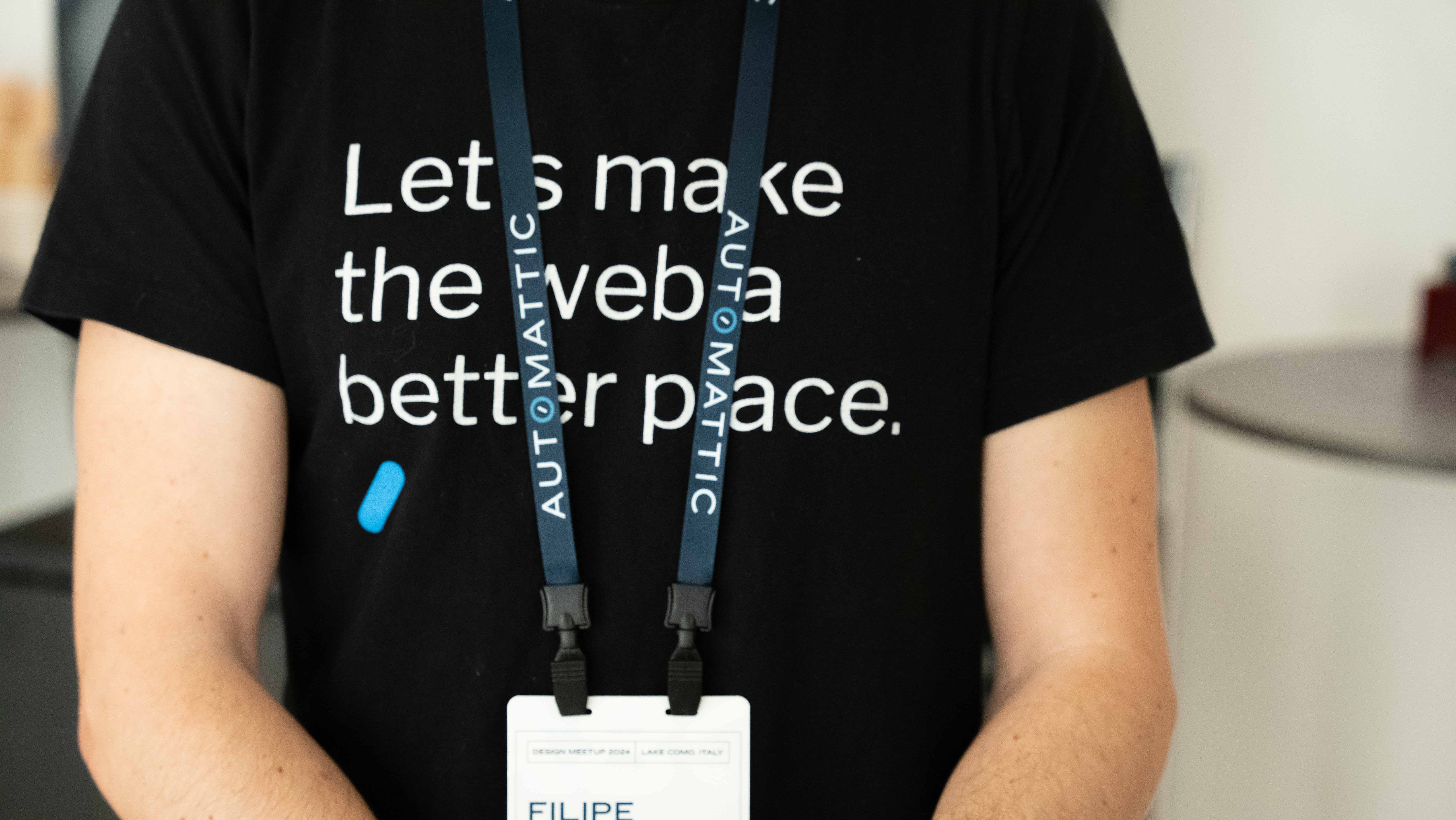 An Automattic employee wearing a black t-shirt with the text 'Let's make the web a better place.' and a lanyard displaying their name and affiliation with Automattic, standing in an informal setting.