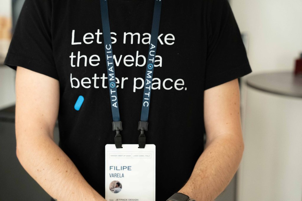 An Automattic employee wearing a black t-shirt with the text 'Let's make the web a better place.' and a lanyard displaying their name and affiliation with Automattic, standing in an informal setting.