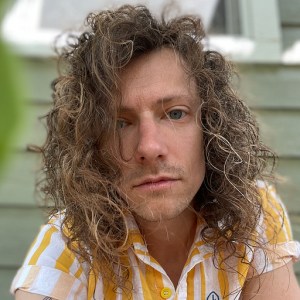 A close-up portrait of a Jerry Janes, an Automattic engineer, wearing a striped shirt, looking directly at the camera with a neutral expression against a background featuring a wooden structure.